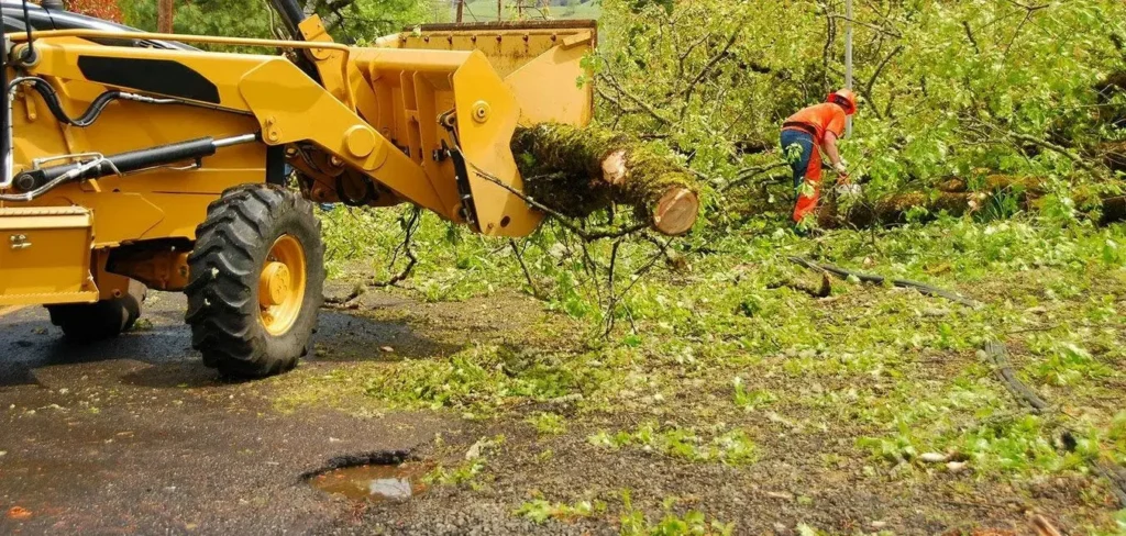 Yellow front-end loader lifting a large mossy log with a worker in orange safety gear standing nearby