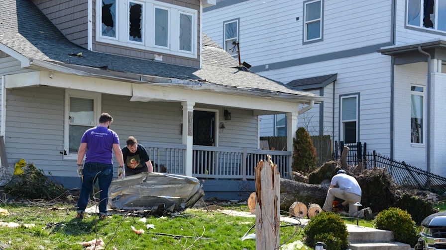 Men clearing debris and cutting a fallen tree in front of a house with storm damage to the roof and windows